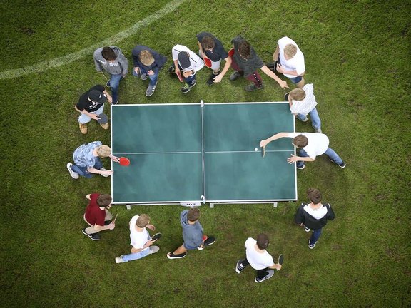 Vogelperspektive auf eine Tischtennisplatte, die auf dem Rasen steht. Drumherum stehen mehrere Menschen mit Schlägern in der Hand an der Platte und spielen Rundlauf. 