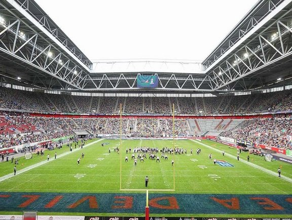 Blick aus dem Rang auf das Football-Feld. Hinter dem Gabelförmigen Football-Tor sieht man die beiden Mannschaften auf dem Feld versammelt. 