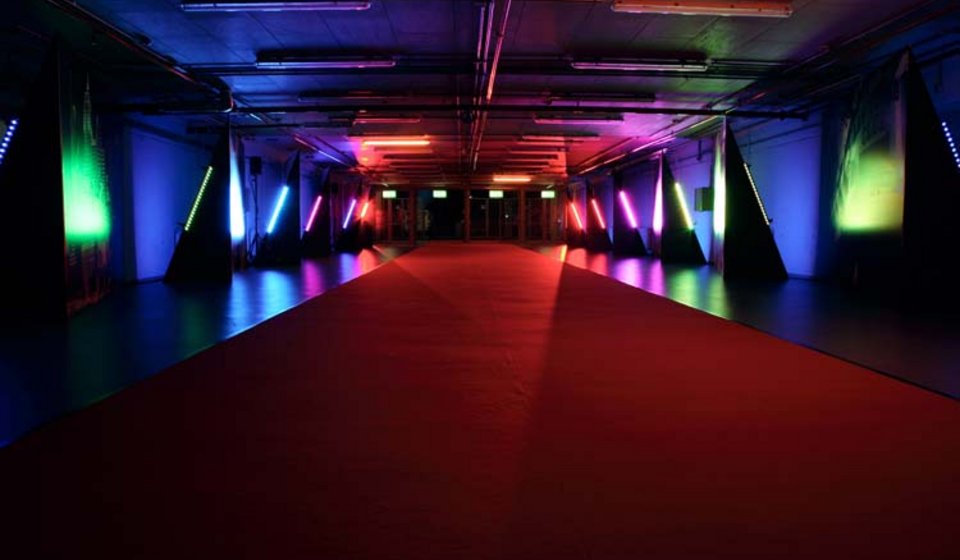 Dunkler Spielertunnel, der durch rote, lilane, blaue und grüne LED-Leisten an den Seiten beleuchtet ist. In der Mitte verläuft ein roter Teppich. 