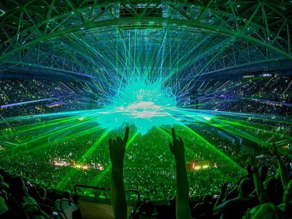 Konzert in der Arena bei geschlossenem Dach. Von der Bühne strahlen grüne und blaue Lichter die Halle an. Zentral werden Zwei Hände in die Luft gehalten. 
