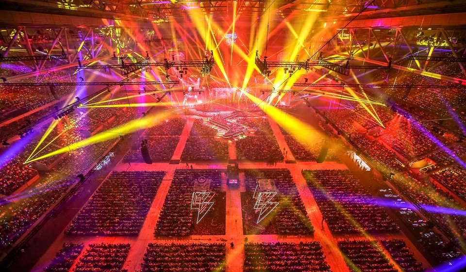 Show in der gefüllten MERKUR SPIEL-ARENA. Über den Köpfen der Zuschauer:innen hängen leuchtende Blitze. Der Innenraum der Arena wird mit gelben und blauen Lichtkegeln angestrahlt. 