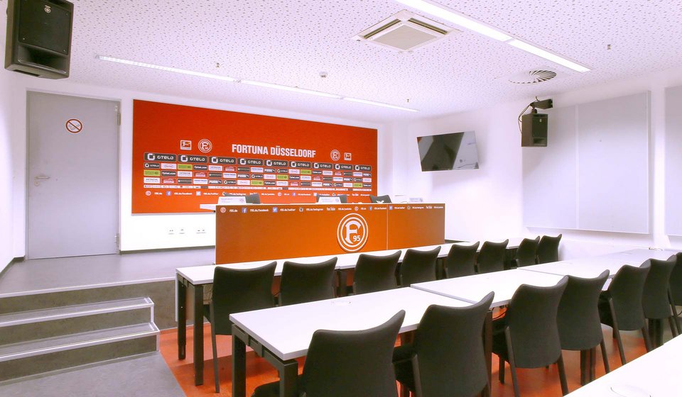Auf einer Bühne sieht man einen Tisch mit Stühlen, der mit einem Fortuna-Düsseldorf-Banner versehen ist. An der Wand dahinter hängt ein Banner von Fortuna mit allen Sponsoren. Vor der Bühne stehen mehrere Tische und Stühle in Reihen. 
