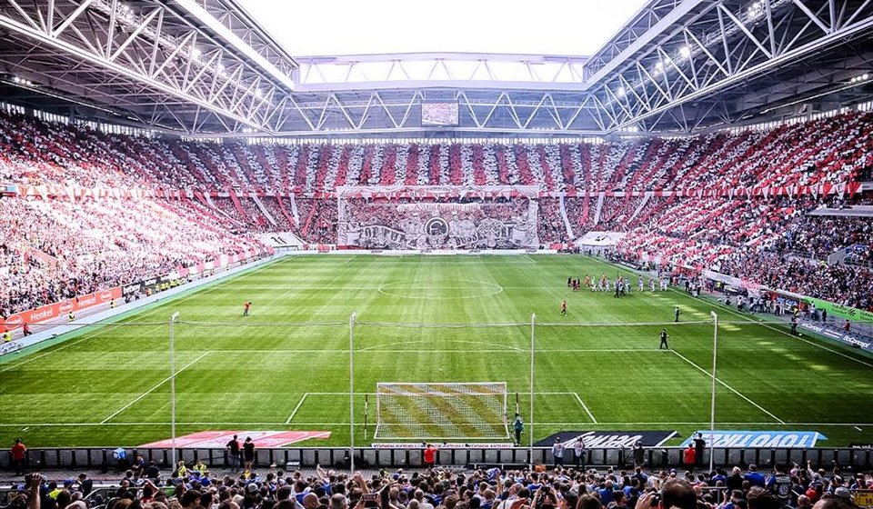 Blick aus dem Rang auf das Fußballfeld bei einem Fortunaspiel in der MERKUR SPIEL-ARENA. Die Fans im Hintergrund sorgen für ein rot-weiß gestreiftes Auftreten der Ränge. 