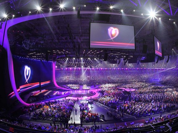 An der Decke der MERKUR SPIEL-ARENA hängen mehrere Übertragungsbildschirme mit dem Logo vom Eurovision Song-Contest. Die gefüllte Halle ist lila beleuchtet. Auf der linken Seite ragt die Bühne in den Innenraum hinein.