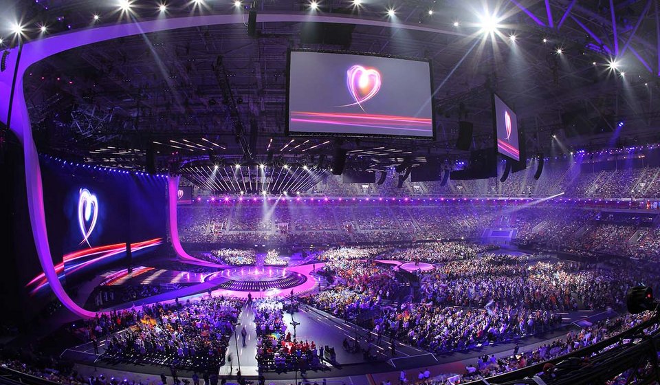An der Decke der MERKUR SPIEL-ARENA hängen mehrere Übertragungsbildschirme mit dem Logo vom Eurovision Song-Contest. Die gefüllte Halle ist lila beleuchtet. Auf der linken Seite ragt die Bühne in den Innenraum hinein.