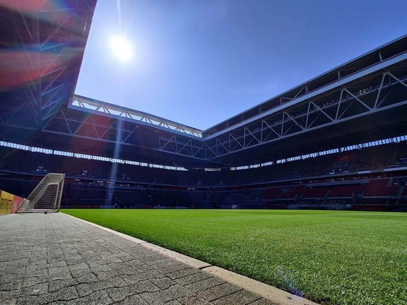 Aufnahme vom Fußballfeldrand. Durch das geöffnete Dach scheint die Sonne auf das Feld. Am Rand sieht man ein Tor stehen. 